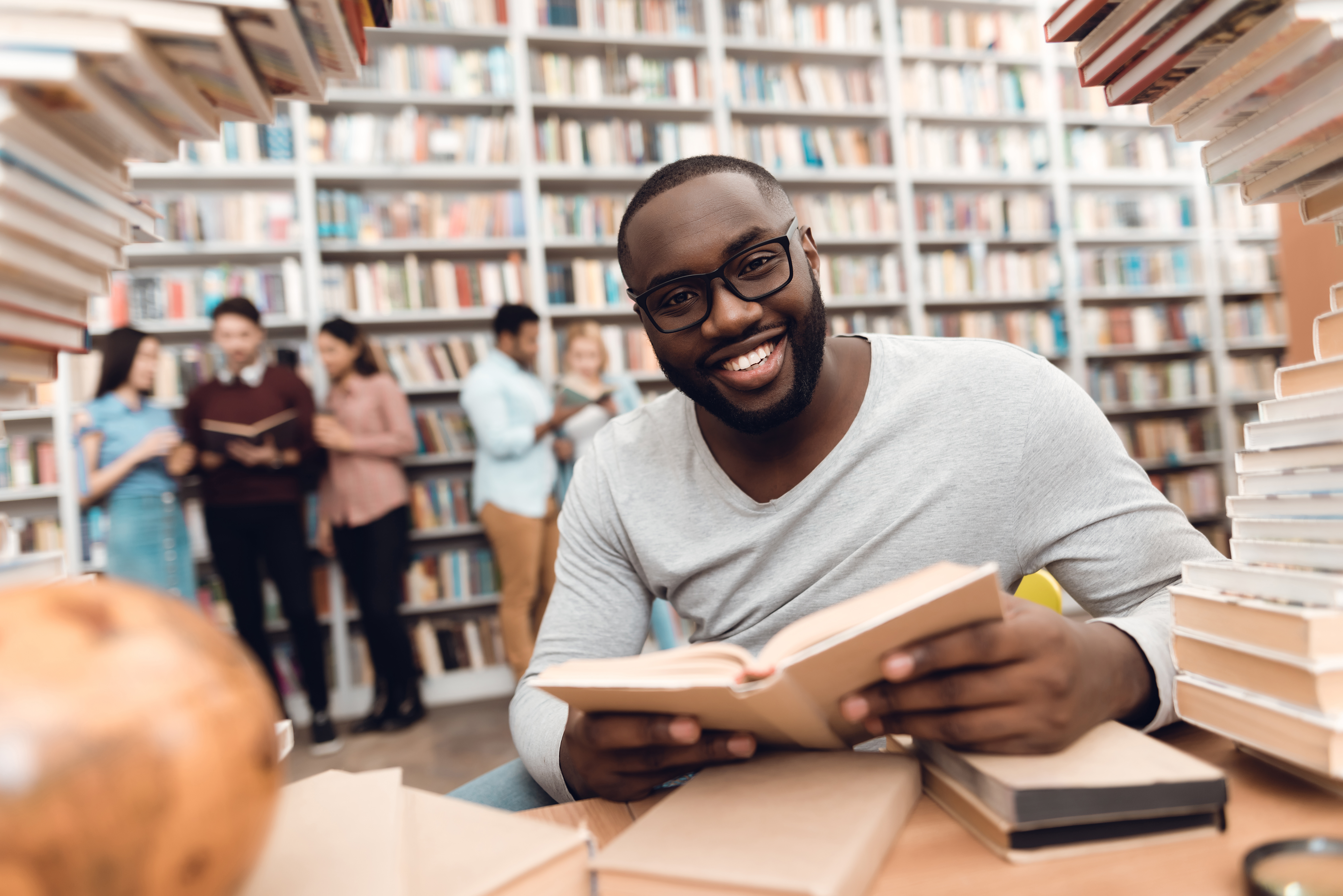 Ethnic african american guy sitting at table surrounded by books in library. Student is reading book.